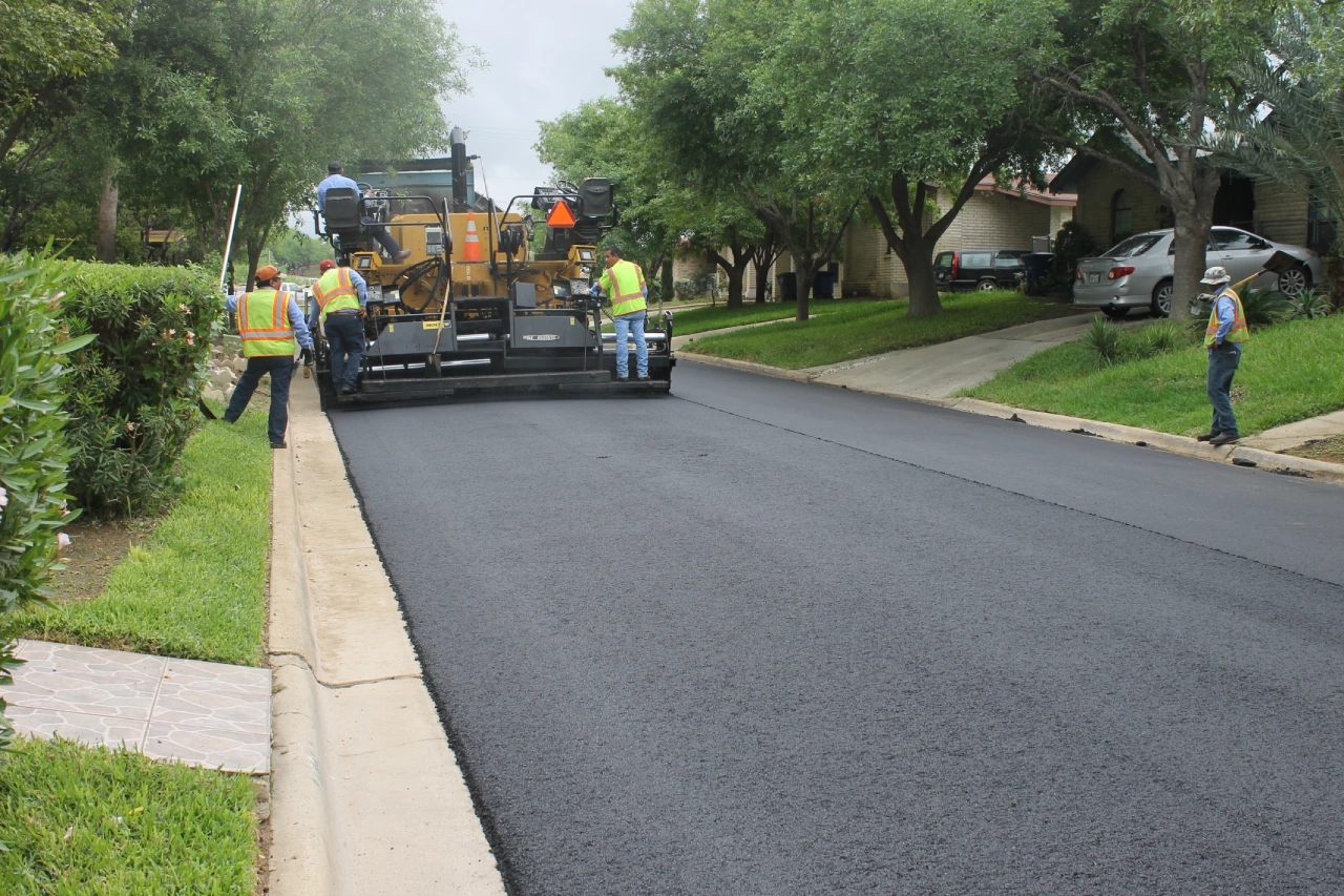 A construction crew operating a heavy asphalt paver during an industrial roadway paving project in a residential neighborhood.